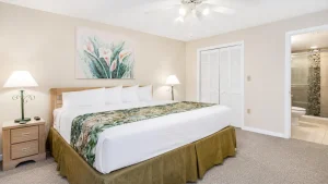 Hotel room with a large bed, white linens, tropical-plant runner, and two bedside lamps on wooden nightstands.