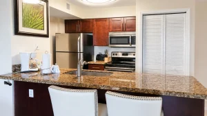 Modern kitchen with granite island, stainless steel fridge, microwave, oven, and dark wood cabinets; white bar stools at the island.