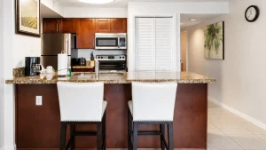 Kitchen island with granite countertop, two white upholstered stools, and stainless steel appliances in a modern open kitchen.