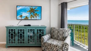 Coastal living room with a turquoise cabinet, wall-mounted TV showing palm trees, and a patterned armchair by a balcony with an ocean view.