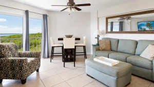 Living room with light blue sofa and patterned armchairs, facing a large window view of greenery and ocean beyond. A dining table and chairs sit to the left, with a ceiling fan above.