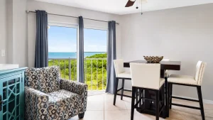 Dining area with a dark wood table and cream upholstered chairs, ocean view visible through large windows with blue curtains.