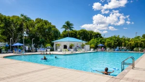 Outdoor resort pool area with palm trees, blue umbrellas, and a blue-roofed lounge building under a sunny sky.
