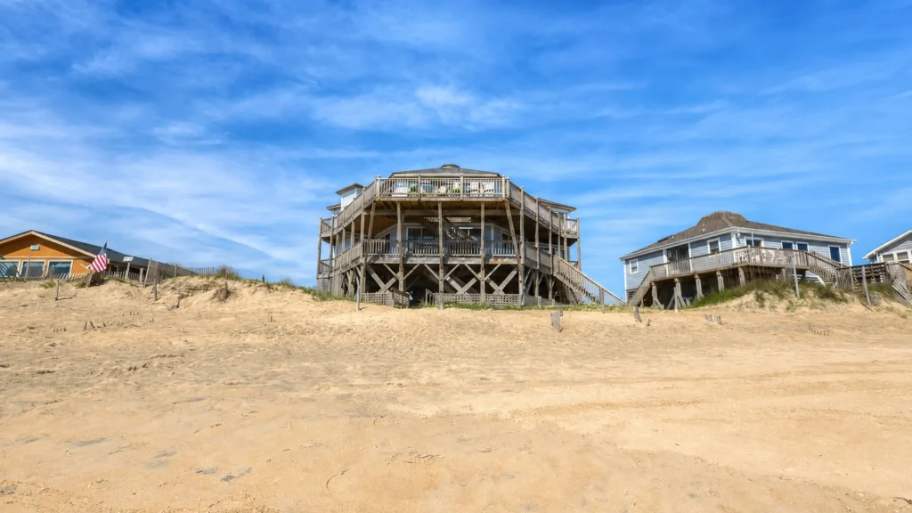 Row of wooden beach houses on stilts above sandy beach under a blue sky.