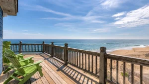 Ocean view from a wooden deck with green Adirondack chairs, railing, and a sandy beach beyond the railing.