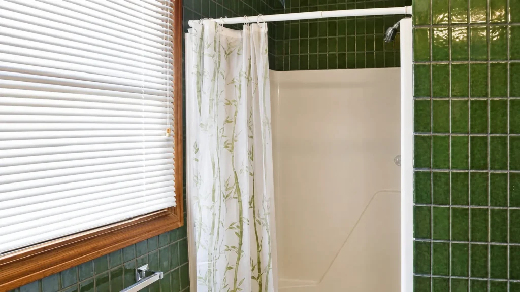 Bathroom shower with white tub, green tile walls, bamboo-pattern shower curtain, and a wooden-framed window with blinds