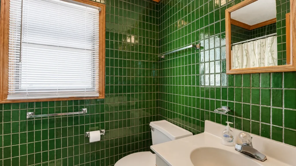 Bathroom with glossy dark-green tiled walls, white sink, toilet, window with white blinds, and a wooden-framed mirror above the sink.