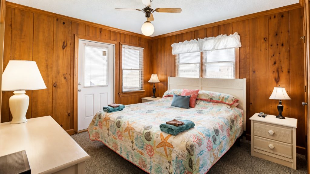 Bedroom with white headboard, starfish quilt, and wood-paneled walls; two nightstands and lamps on either side of the bed, window blinds behind.