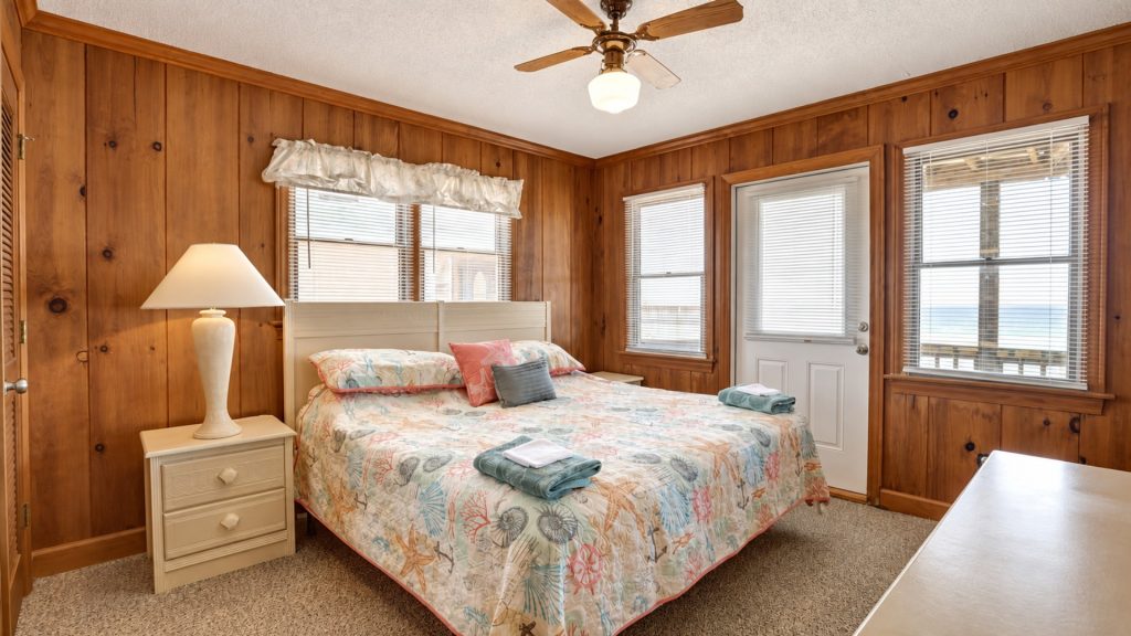 Cozy beach-themed bedroom with wood-paneled walls, queen bed, white nightstand, and ocean view through the windows.
