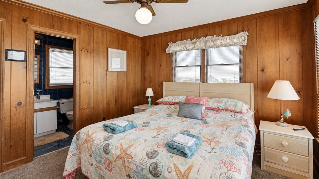 Wood-paneled bedroom with a seashell-patterned quilt on a white bed, matching nightstands, and a window with blinds; doorway reveals a blue-tiled bathroom.