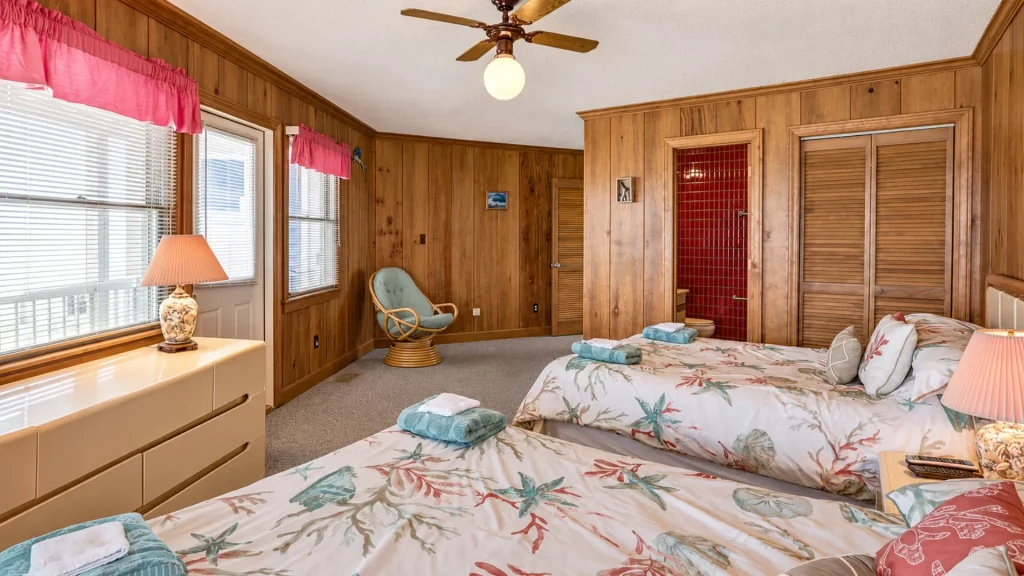 Bedroom with wood panel walls, two beds with tropical bedding, teal towels, and a red-tiled bathroom visible in an alcove.
