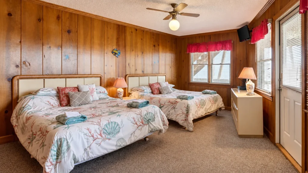 Coastal-themed hotel room with two queen beds, seashell bedding, and wood-paneled walls.