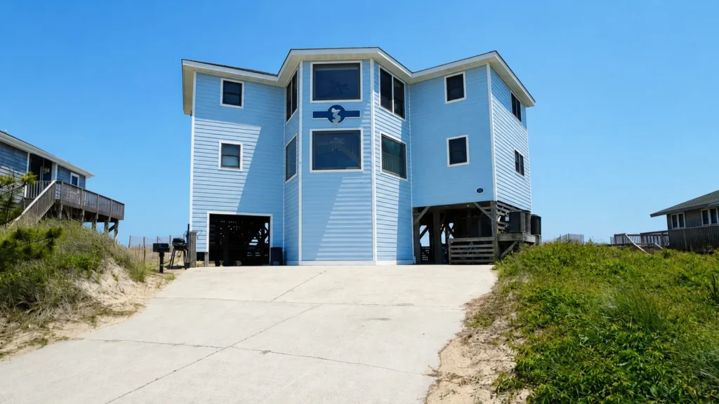 Blue beach house on stilts with large windows, raised above sandy dunes under a clear blue sky.