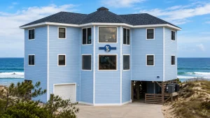 Blue, three-story beach house on stilts with white trim and a large central bay window overlooking the ocean.