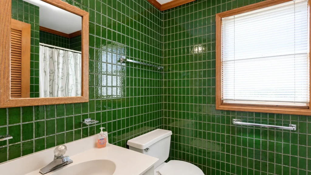 Green tiled bathroom with a wooden-framed mirror, window with blinds, and white sink and toilet fixtures.