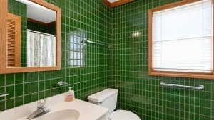 pompous-pelican-full-bath-1-1 | Swack Vacations Green tiled bathroom with a wooden-framed mirror, window with blinds, and white sink and toilet fixtures.