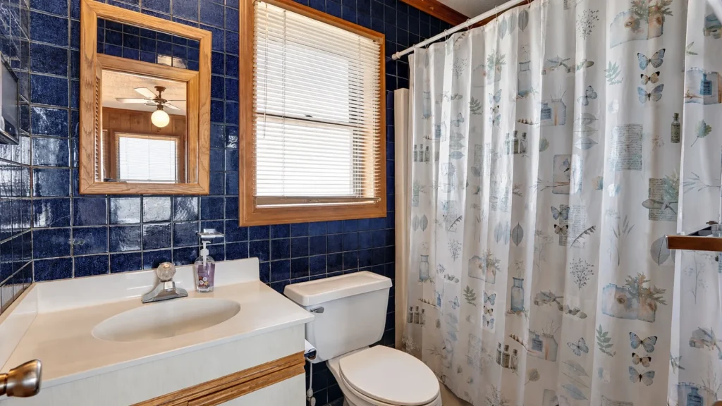 Bathroom with dark blue tiled walls, a wood-framed mirror above a white sink, and a white toilet beside a butterfly-pattern shower curtain.