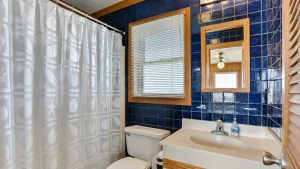 pompous-pelican-full-bath-3-1 | Swack Vacations Blue tiled bathroom with a white sink and toilet, a seashell-patterned shower curtain, and a window with horizontal blinds framed by wood trim.