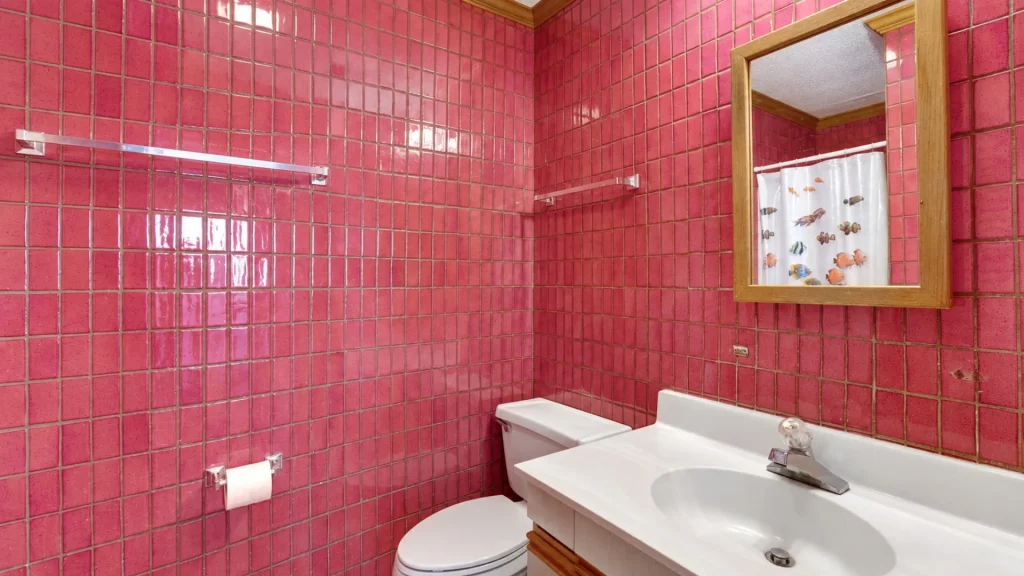Pink-tiled bathroom with a white sink and toilet, wooden-framed mirror, and a fish-themed shower curtain reflected in the mirror.