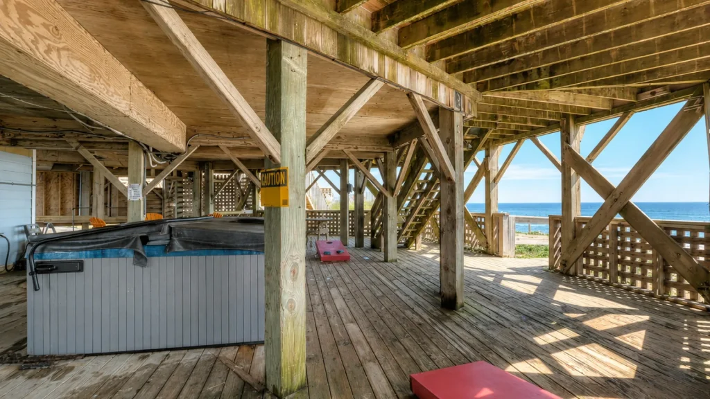 Under a coastal wooden deck with weathered beams; a hot tub on the left, a yellow caution sign on a post, and the ocean visible in the background.