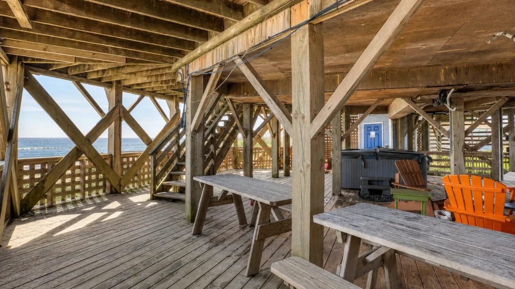 Wooden beach deck with picnic tables and colorful Adirondack chairs, overlooking the ocean, under a weathered wooden structure with diagonal support beams.