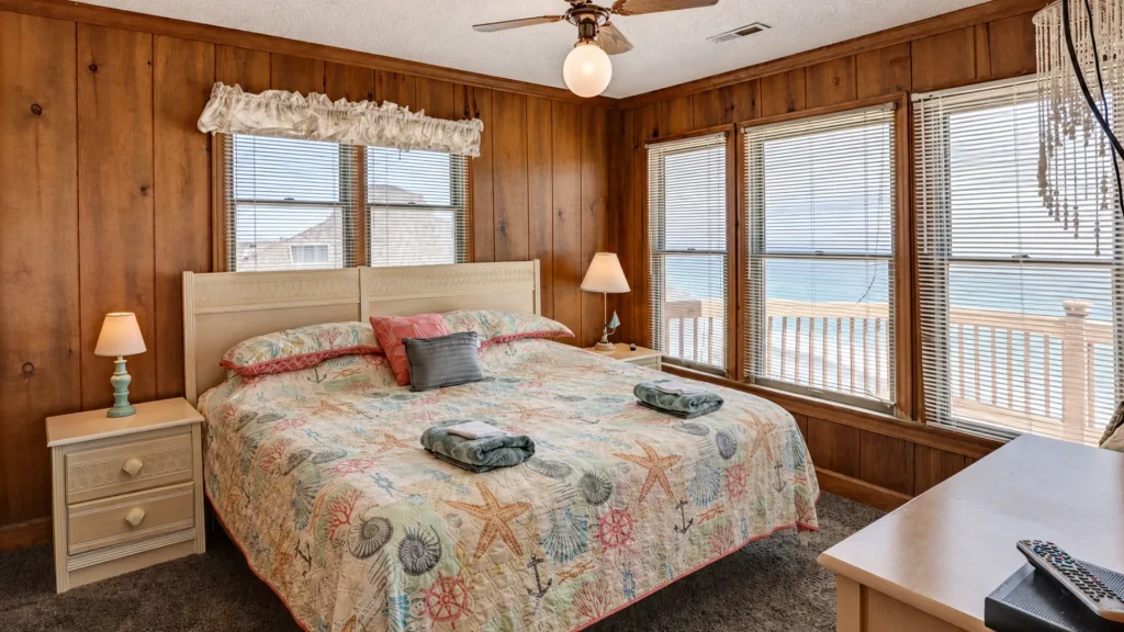 Coastal bedroom with wood-paneled walls, white bed, seashell quilt, and large windows with blinds overlooking the ocean.