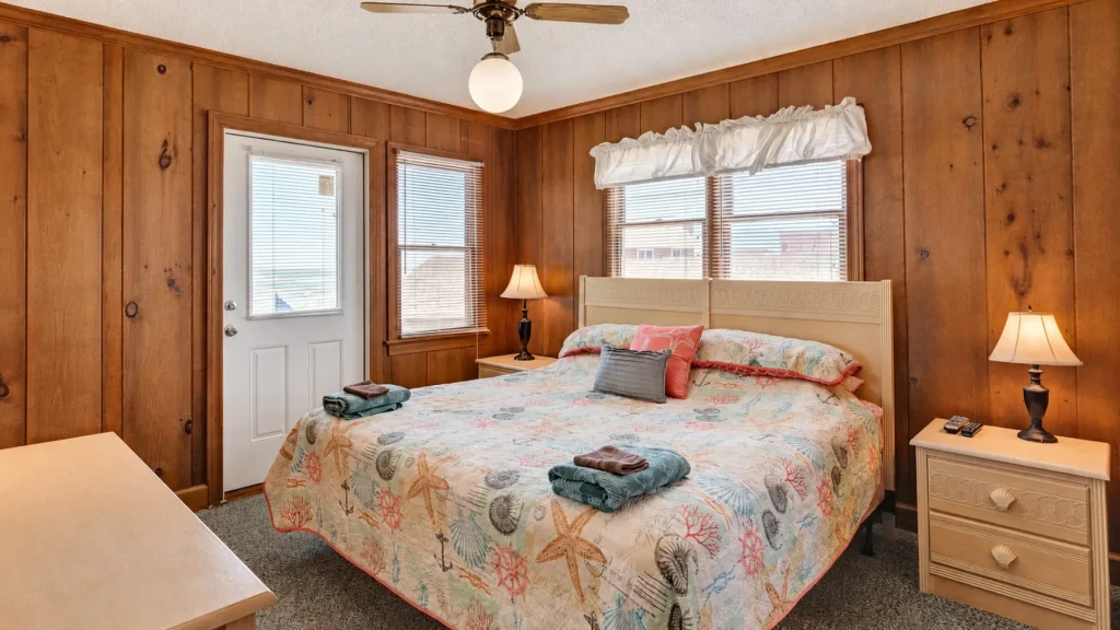 Wood-paneled bedroom with a white bed, sea-themed quilt, and folded towels on the foot of the bed, two nightstands with lamps, and ceiling fan.