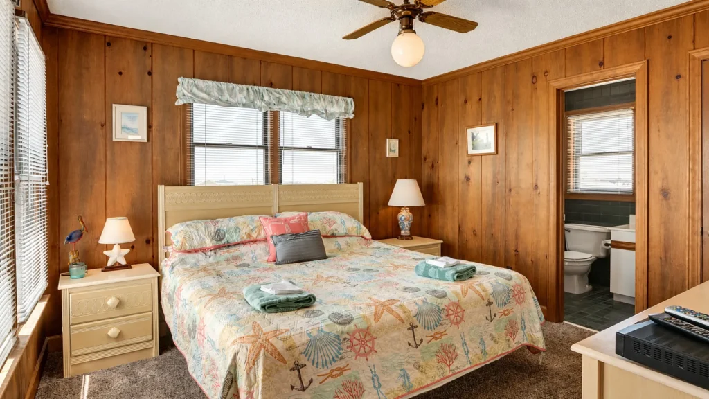 Bedroom with wood-paneled walls, a queen bed in sea-themed quilt, two cream nightstands, and a doorway to a bathroom.