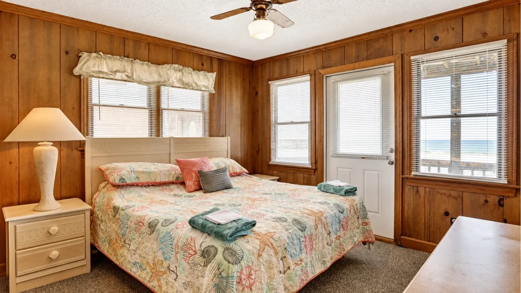 Cozy coastal bedroom with a sea-themed quilt, pastel pillows, and wood-paneled walls, with an ocean view through the windows.