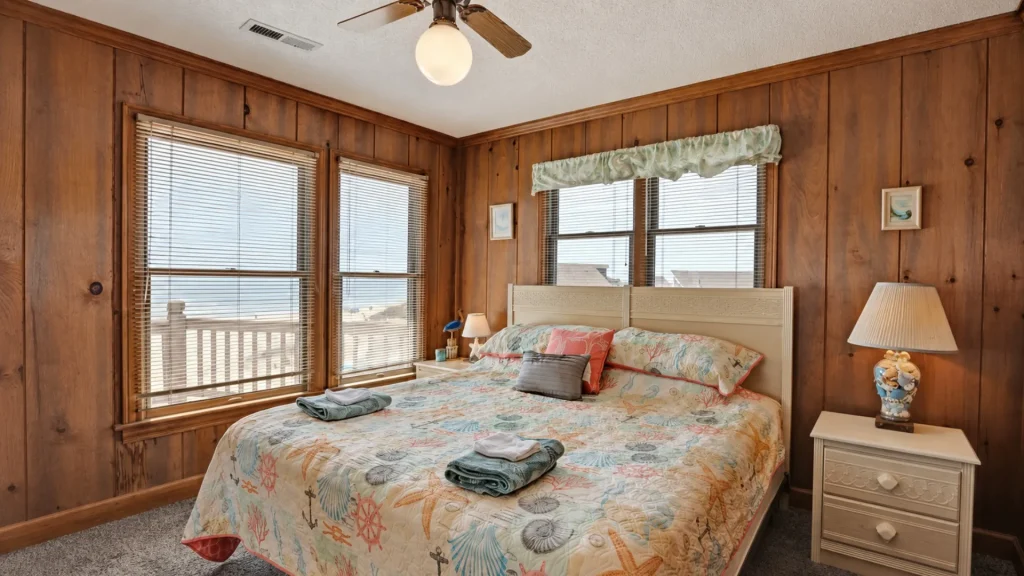 Coastal bedroom with wood paneled walls, a cream headboard bed, and large windows with blinds overlooking the sea.