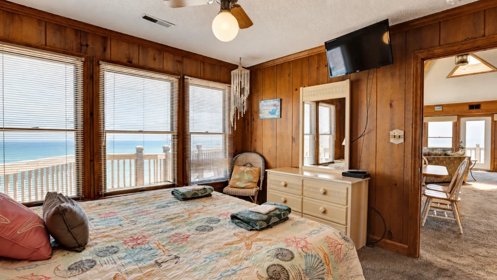 Bedroom with wood-panel walls and large windows showing an ocean view; shell-pattern quilt on the bed and a dresser with a mirror nearby.