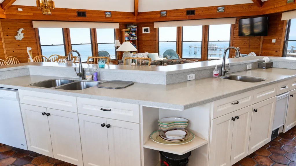 Open kitchen with white cabinets and a long island, showing a living area and ocean view through bay windows.