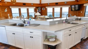 Open kitchen with white cabinets and a long island, showing a living area and ocean view through bay windows.
