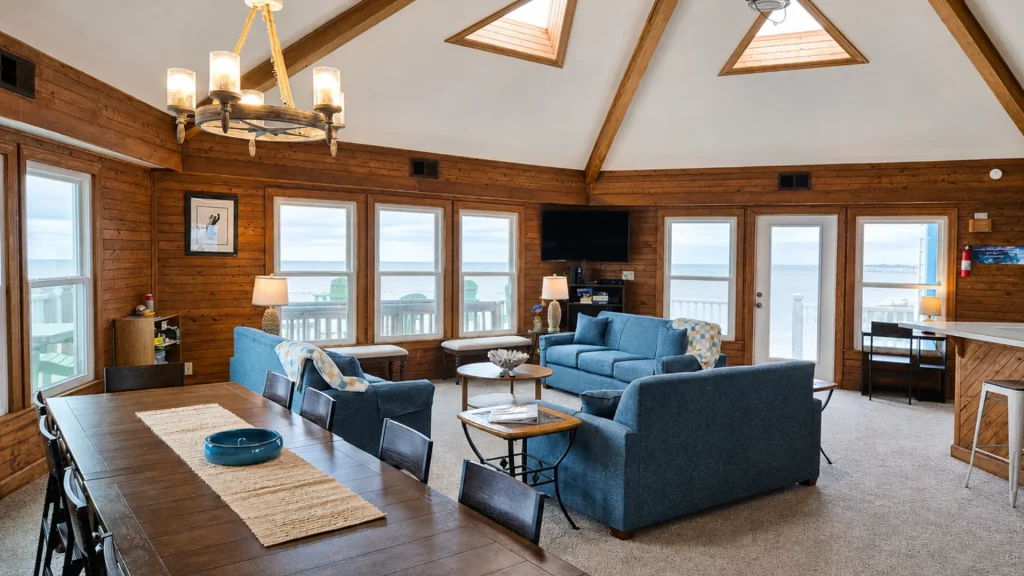 Cozy coastal living room with wood-paneled walls, blue sofas, and a vaulted ceiling with skylights overlooking the ocean outside windowed wall