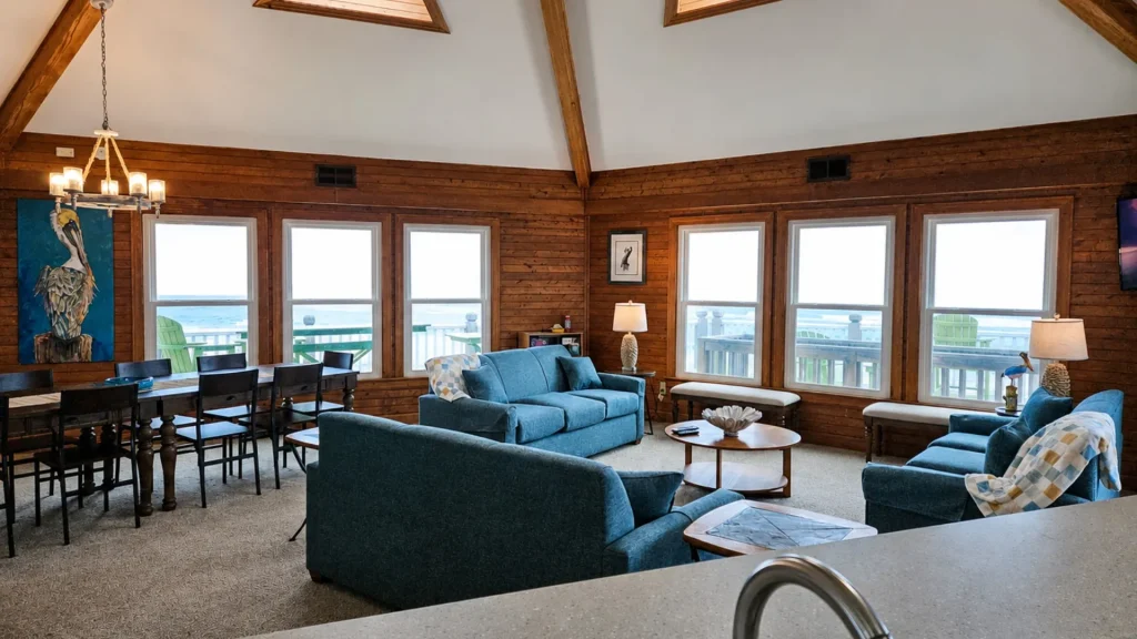 Cozy lakefront living room with blue sofas, wooden walls and vaulted ceiling, large windows showing a water view and deck outside, plus a dining table on the left.