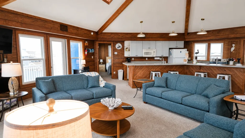 Open living area with blue sofas, wood-paneled walls and exposed beams, plus a white-kitchen bar and stools. Large windows/blue door visible.