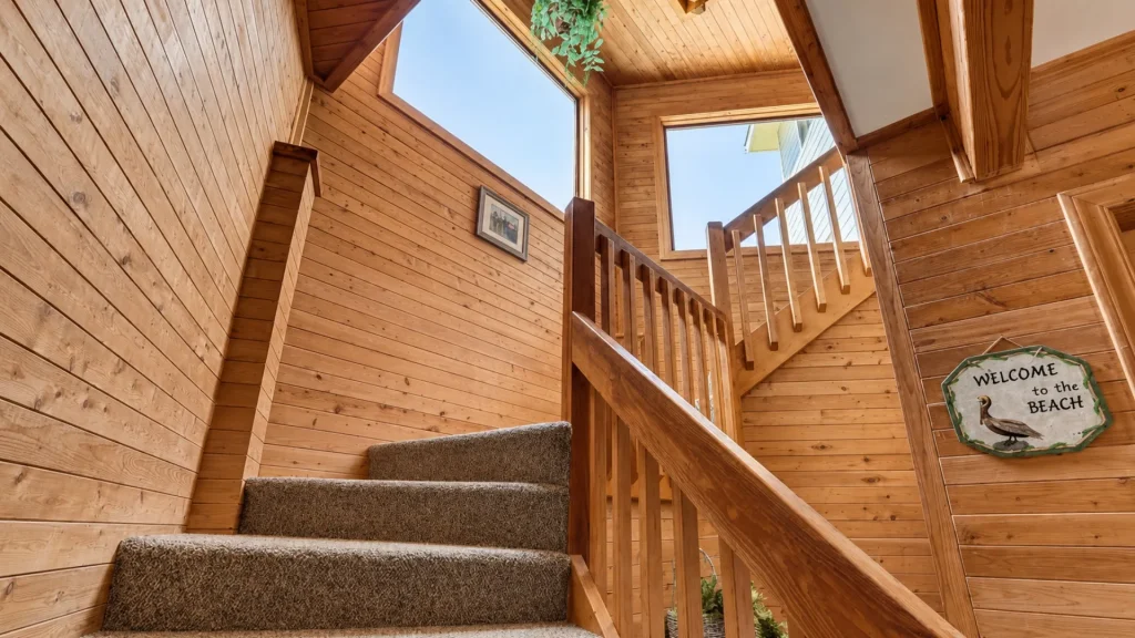 Wood-paneled stairwell with carpeted steps, wooden railing, and large upper windows; beach sign on wall.