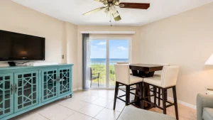 Living room with a turquoise cabinet, wall-mounted TV, and a sliding glass door opening to a balcony with an ocean view.