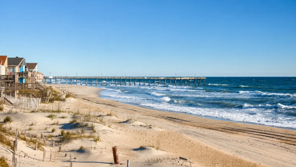 Beache front houses along a sandy shore with a wooden pier extending into the blue ocean under a clear sky, left side of image.