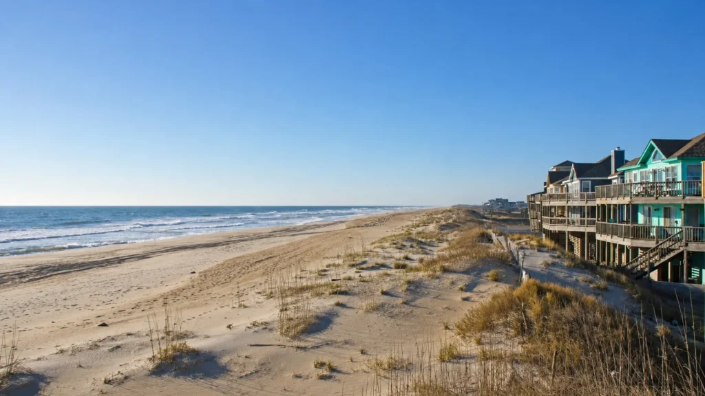 Row of colorful beach houses on stilts along a sandy dune, with the ocean to the left.