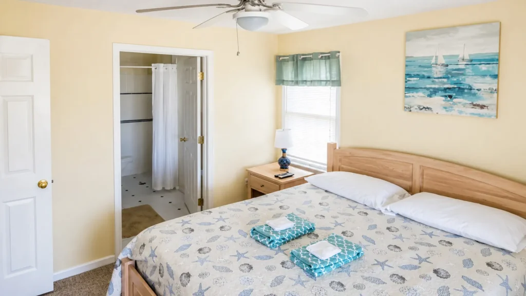 Cozy bedroom with a sea-themed quilt, two pillows, and a wooden bed; nightstand lamp, ocean painting, and a window with a teal valance on pale yellow walls; doorway to a bathroom with a white shower curtain visible.