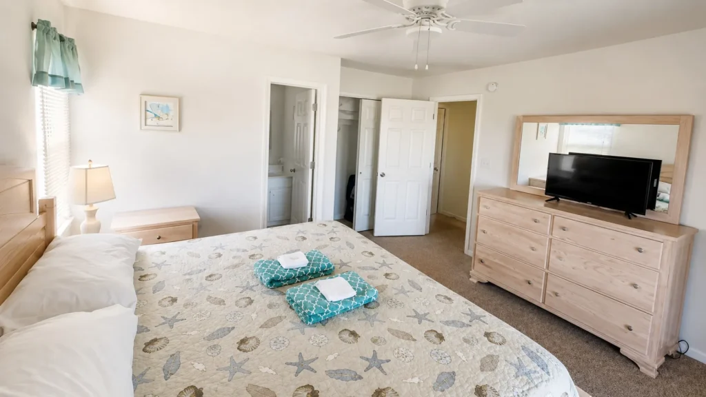 Bright hotel bedroom with a sea-themed quilt, two turquoise folded towels on the bed, and a dresser with a TV and mirror.