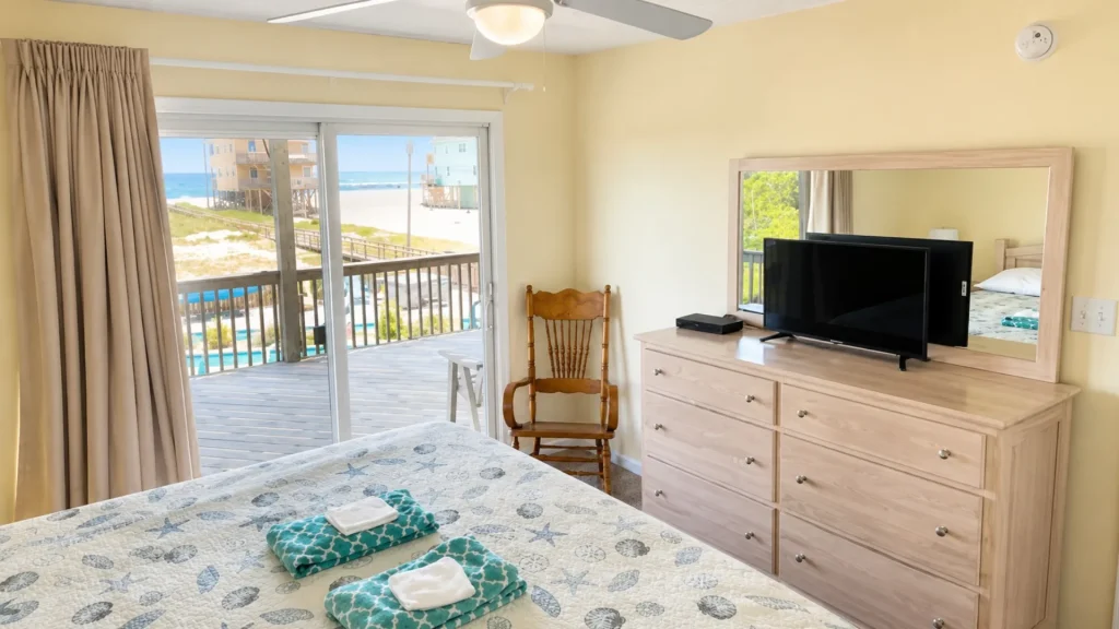 Bedroom in a beachside hotel with a sliding glass door to a balcony overlooking the ocean; dresser with a flat-screen TV and a large mirror above it.