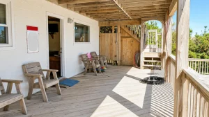 Wooden back deck attached to a white house with several Adirondack chairs, an open door, and wooden stairs in bright sunlight.
