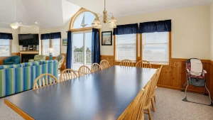 Open family room with a long black dining table and wooden chairs, blue curtains, and a balcony door at the back sunlit by large windows.