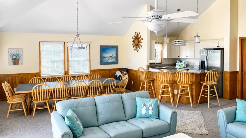 Open living space with a blue sofa, teal pillows, and a long wooden dining table with chairs; kitchen visible in the background.