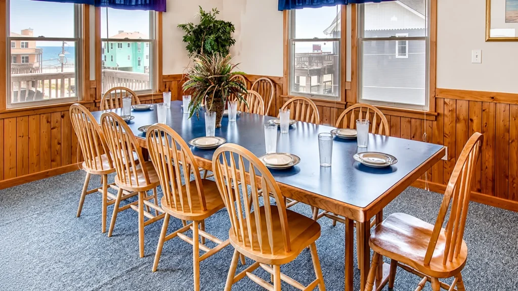Long blue dining table set for a meal in a sunlit coastal dining room with wooden panel walls and windows overlooking the beach town view.