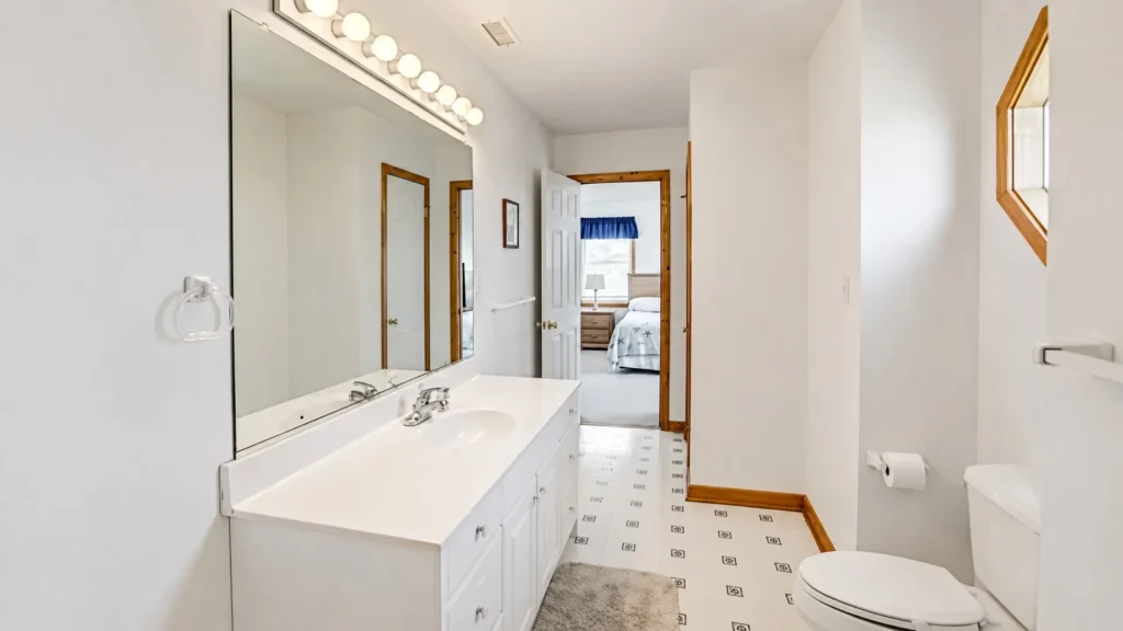 White bathroom with a double-sink vanity, large mirror, and row of globe lights above; doorway opens to a bedroom with a blue valance.