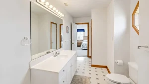 White bathroom with a double-sink vanity, large mirror, and row of globe lights above; doorway opens to a bedroom with a blue valance.