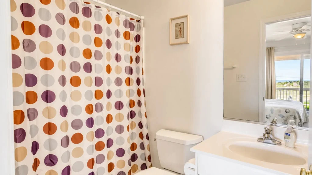Bright bathroom with a colorful polka-dot shower curtain, white toilet, and sink; mirror reflects a bedroom with a balcony view outside.